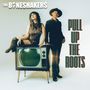 "THE BONESHAKERS" oben links, "PULL UP THE ROOTS" rechts; Frau auf Vintage-TV, Mann steht daneben in dunkler Kleidung.