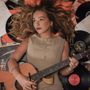 Eine Frau mit lockigem Haar liegt auf Schallplatten und hält eine Gitarre. Ein Notenblatt ist im Hintergrund sichtbar.