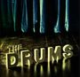 Text: "THE DRUMS". Große 3D-Buchstaben auf einem Holzboden, im Hintergrund ein schwerer, blauer Vorhang.