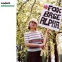 Text: „FOX BASE ALPHA“ auf einem Schild. Frau in gestreiftem Shirt, Hut, hält Schild vor Bäumen. Logo: „saint etienne“.