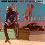 Oben steht "SEU JORGE THE OTHER SIDE". Ein Mann mit Hut sitzt auf einem Container vor einer bemalten Wand.