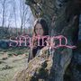 Text: "SMEKKIÐ" in pinker Schrift. Eine Person steht hinter einem Baum in einer Waldlandschaft.