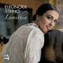 "Eleonora Strino, Lumithia." Eine Frau mit dunklen Haaren lächelt und hält eine Gitarre, trägt ein weißes Hemd.
