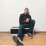 Eine Person sitzt entspannt in Jeans und Mütze vor einer weißen Backsteinwand auf einem Kasten.