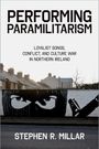 "PERFORMING PARAMILITARISM" zeigt Häuser und ein Wandgemälde mit Augen und einer vermummten Person.