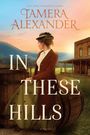 Text: "Tamera Alexander. In These Hills." Eine Frau im historischen Outfit steht neben einem Fass vor einem Holzgebäude.