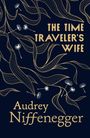 „The Time Traveler's Wife“ und „Audrey Niffenegger“ in goldener Schrift. Design mit goldenen Wellenlinien und weißen Pusteblumen.