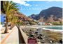 "LA GOMERA", "20x7 CASAES EDITION". Strandpromenade mit Palmen, Meer, schwarzer Lavasteinküste, Berge im Hintergrund.