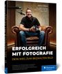 „Erfolgreich mit Fotografie: Dein Weg zum bezahlten Bild.“ Mann mit Kamera sitzt lächelnd auf einem Ledersessel.