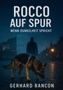 "ROCCO AUF SPUR - WENN DUNKELHEIT SPRICHT" von Gerhard Rancon. Ein Hund auf Kopfsteinpflaster, Silhouette eines Mannes im Hintergrund.