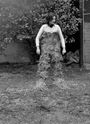 Keine sichtbaren Texte. Eine Person in einem Garten trägt Heu als Kleidungsstück. Im Hintergrund sind Pflanzen zu sehen.