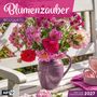 „Blumenzauber Bouquets“, ein Strauß bunter Blumen in einer violetten Vase auf einer weißen Bank.