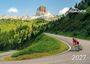 "2027" und "QUÄLDICH" Logo. Radfahrer in rotem Trikot windet sich an grüner Bergstraße entlang. Bergkulisse im Hintergrund.