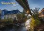 "Wuppertal 2027" steht groß im Bild. Eine Schwebebahn fährt über einen Fluss, daneben alte Gebäude und Bäume im Herbst.