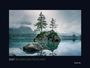 2027 WILDNIS DEUTSCHLAND. Ein stiller See mit Bäumen auf einem Felsen und nebelverhangenen Bergen im Hintergrund.