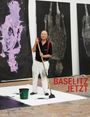 "BASELITZ JETZT" in roten Buchstaben. Ein älterer Mann steht mit einem großen Pinsel auf einem Atelierboden.